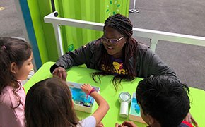 A group of children is engaged in a hands-on science activity at a bright green table, where they are exploring a water filtration experiment. The facilitator, wearing a SPARK™ shirt, demonstrates the process while the children attentively observe and participate, fostering an interactive learning experience outdoors.