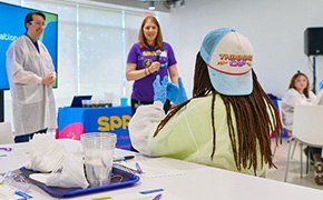A child wearing a "Thinking Cap" hat is seated at a table, facing a presentation about water filtration, with two adults standing in front of a large screen displaying relevant information. The adults are engaged in explaining the topic, while various educational materials, including cups and worksheets, are visible on the table, creating an interactive learning environment.