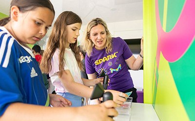 Three children are engaged in an interactive activity at a colorful educational booth, with one child holding a device while another observes intently. An adult, wearing a purple SPARK™ volunteer shirt, is guiding them through the activity, promoting hands-on learning and engagement. The vibrant background features bright colors and patterns, enhancing the lively atmosphere of the event.