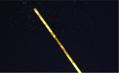 Photo of Overcoated SPME Fiber After Repeated Extractions An image of a long, thin, yellowish rod or stick against a dark background. The rod appears to be illuminated, highlighting its surface texture, which shows some irregularities. Small particles or specks of light are visible around the rod, suggesting either dust or a mist in the air, creating a sense of depth in the image. The overall composition emphasizes the rod's length and the contrast between its color and the dark backdrop.