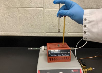 Spiking of Carbotrap® T420 tube with the terpene test mix using Supelco® Adsorbent Tube Injector System (ATIS). Right Hand wearing a blue glove shown adding terpene test mix using Supelco® Adsorbent Tube Injector System (ATIS) to the Carbotrap® T420 tube kept on a platform against a white colored wall.