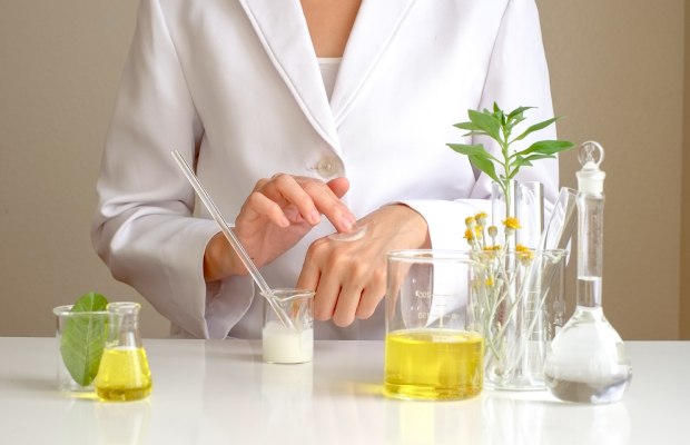 Fragrance Safety in Cosmetics Scientist in a lab coat rubs white cream on the back of their hand with one finger.