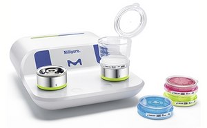 Un sistema de filtración microbiana Millipore con dos unidades de filtración arriba y tres placas de Petri con muestras cultivadas delante. El sistema es blanco y azul con el logotipo de Millipore. Las placas de Petri contienen medios de colores: azul, rosa y amarillo.
