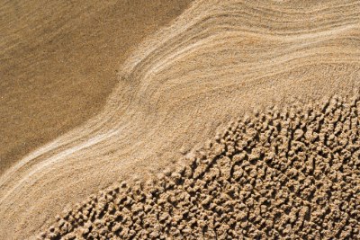 Sand and particulate matter A close-up view of a sand, featuring textured patterns of wet and dry sand, with gentle waves creating smooth lines on the surface