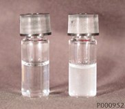 Precipitation of Lipophilic Components of Hydrocortisone Cream When Exposed to DI H2O:Methanol (1:1). (left vial- blank; right vial- precipitated sample) Two small glass vials on a neutral background. The vial on the left contains a clear liquid, while the vial on the right holds a cloudy, white liquid, both sealed with silver caps.