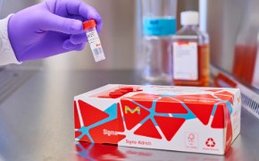 A gloved hand reaches into the tissue culture hood from the top left side of the image. It is holding a MilliShot vial that it has removed from the open box of product in the foreground. The box has vials in it, with the caps visible. In the background, on the right, is a bottle of cell culture media next to a Stericup filter. A gloved hand reaches into the tissue culture hood from the top left side of the image. It is holding a MilliShot vial that it has removed from the open box of product in the foreground. The box has vials in it, with the caps visible. In the background, on the right, is a bottle of cell culture media next to a Stericup filter.